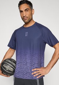 Αθλητικό μπλουζάκι ναυτικού μπλε με διαβαθμισμένο μοτίβο σε μωβ αποχρώσεις, κοντά μανίκια και λογότυπο Under Armour. Το μοντέλο κρατάει μια μαύρη μπάλα μπάσκετ Spalding.