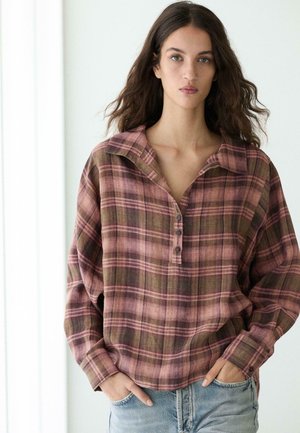 Žena s dlouhými vlnitými vlasy, která nosí volnou růžovo-hnědou kostkovanou košili na knoflíky a světle modré džíny, stojící s rukama v kapsách.