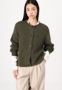 Cardigan tricotat de culoare verde închis, cu guler rotund, mâneci lungi și cinci nasturi negri în față. Textură moale, croi ușor supradimensionat.