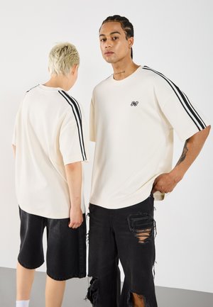 T-shirts crème surdimensionnés avec des rayures noires sur les manches, arborant un petit logo. Des modèles portent des shorts noirs déchirés, se tenant dos à dos.
