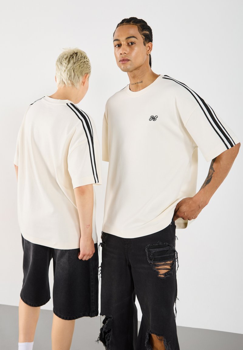 Overmaatse crème t-shirts met zwarte strepen op de mouwen, voorzien van een klein logo. Modellen dragen versleten zwarte shorts en staan rug aan rug.