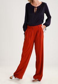 Blouse marine avec un col à nouer en forme de clé, associée à un pantalon taille haute plissé de couleur rouge vif. Modèle portant des sandales métalliques.
