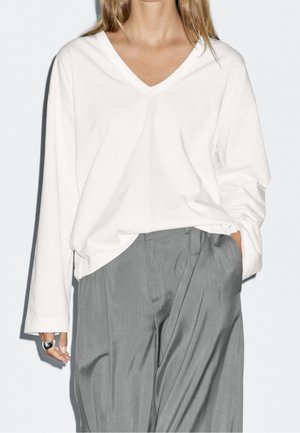 Woman with long hair wearing a white V-neck long-sleeve top tucked into high-waisted gray wide-leg pants, hand in pocket.