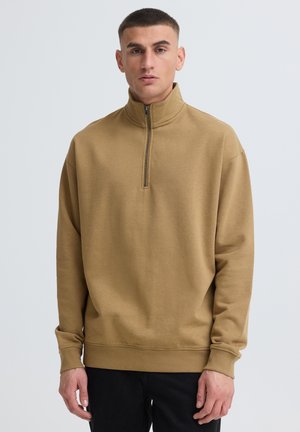 Ung mand med kort hår iført en beige lynlås-trøje og sorte bukser mod en ensfarvet lys baggrund.