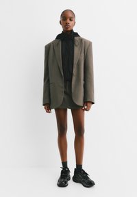 Blazer marron surdimensionné sur une veste à capuche noire, associé à une mini-jupe et des baskets noires. Texture lisse, design structuré.