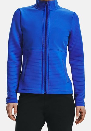 Vrouw in een koningsblauwe zip-up jack met horizontale geribbelde details en zwarte broeken, staand met ontspannen armen langs haar zij.