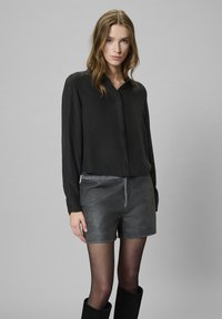 Młoda kobieta ubrana w czarną bluzkę z długim rękawem, szare skórzane szorty, czarne przezroczyste rajstopy i czarne botki, stoi na jednolitym szarym tle.