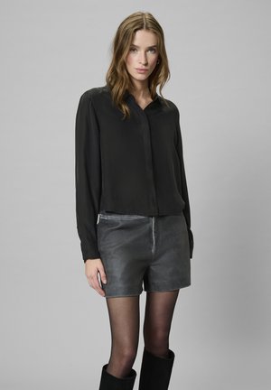 Junge Frau in einer schwarzen Bluse mit langen Ärmeln, grauen Lederhosen, schwarzen, transparenten Strumpfhosen und schwarzen Stiefeln, die vor einem schlichten grauen Hintergrund steht.