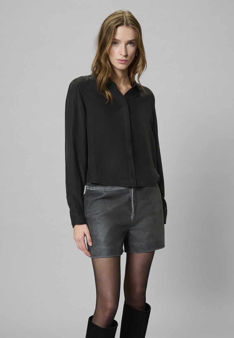 Młoda kobieta ubrana w czarną bluzkę z długim rękawem, szare skórzane szorty, czarne przezroczyste rajstopy i czarne botki, stoi na jednolitym szarym tle.