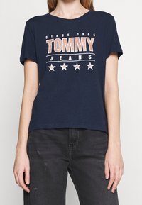 Námořnická modrá bavlněná tričko s nápisem "TOMMY JEANS", hvězdami a designovými prvky "SINCE 1985" v kontrastní bílé a červené barvě. Krátké rukávy, volný střih.