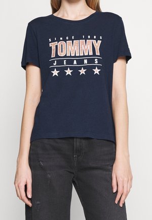 Camiseta de algodón azul marino con el texto "TOMMY JEANS", estrellas y elementos de diseño "SINCE 1985" en colores blanco y rojo contrastantes. Manga corta, corte relajado.