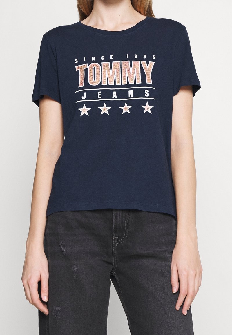 Námořnická modrá bavlněná tričko s nápisem "TOMMY JEANS", hvězdami a designovými prvky "SINCE 1985" v kontrastní bílé a červené barvě. Krátké rukávy, volný střih.