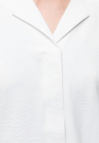 Gros plan d'un vêtement blanc avec un col en V et une patte de boutons cachée sur un ton de peau clair.