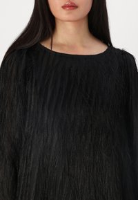 Top nero e testurizzato con lunghe e sottili strisce verticali. Collo rotondo, tessuto leggero, texture liscia e una leggera lucentezza.