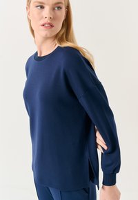 Maglia a maniche lunghe blu navy con scollatura rotonda, spacchi laterali, polsini a coste e una texture liscia. Tessuto morbido con una vestibilità comoda.