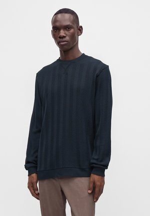 Jeune homme portant un pull bleu foncé texturé à manches longues et un pantalon marron clair, debout devant un fond clair uni.
