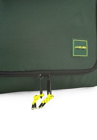 Bolsa de tela texturizada verde con una cremallera negra. Presenta tiradores amarillos y negros y una etiqueta verde y amarilla que dice "JASLEN EST. 1987".