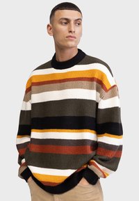 Pull en maille épaisse avec des rayures horizontales en olive, blanc, noir, orange et marron. Encolure et poignets côtelés, coupe décontractée.