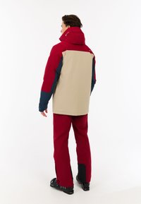 Beige, rote und marineblaue gepolsterte Jacke mit Kapuze, geeignet für den Außenbereich. Trägt dazu passende rote Hose und schwarze Skischuhe.