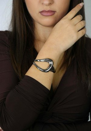 Bracelet en métal argenté incurvé avec un design ouvert, présentant une finition polie et une texture lisse, porté au poignet d'une personne.