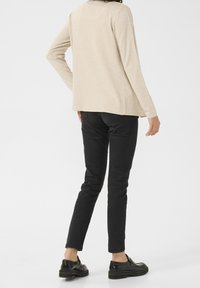 Kobieta w beżowej bluzce z długim rękawem, czarnych wąskich spodniach i czarnych loafersach, stoi plecami z jedną ręką na biodrze na białym tle.
