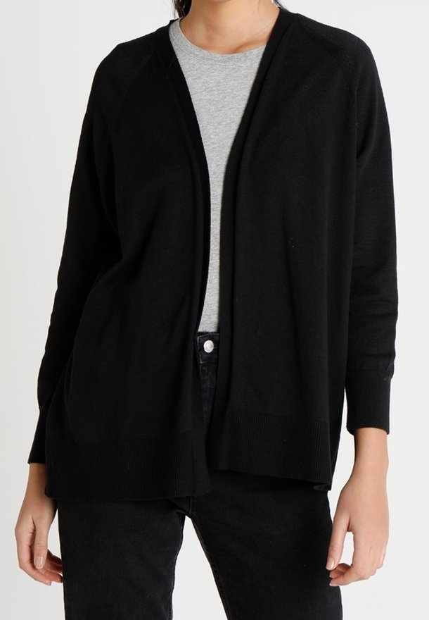 Femme portant un cardigan noir ouvert sur un t-shirt gris à col rond et un pantalon noir, debout devant un fond blanc.