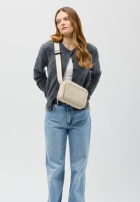 Borsa a tracolla beige in pelle liscia con forma rettangolare, chiusura con zip e tracolla regolabile, indossata con un cardigan grigio e jeans blu.