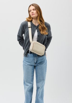 Bolso cruzado beige hecho de cuero suave, con forma rectangular, cierre de cremallera y correa ajustable, combinado con un cárdigan gris y jeans azules.