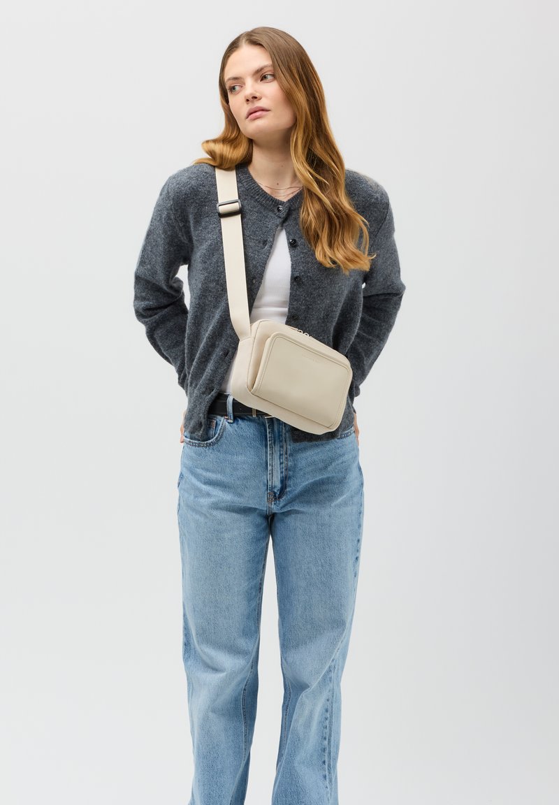 Borsa a tracolla beige in pelle liscia con forma rettangolare, chiusura con zip e tracolla regolabile, indossata con un cardigan grigio e jeans blu.