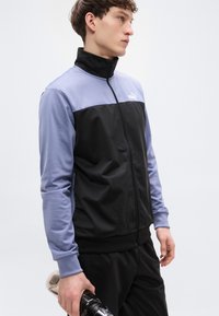 Μαύρο και ανοιχτό μωβ ζακέτα αθλητική με φερμουάρ και ψηλό γιακά, με ελαστικά μανσέτα και λογότυπο στο στήθος. Λείο ύφασμα.