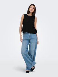 Woman wearing a sleeveless black top with side ties, wide-leg blue jeans, and black pointed shoes, standing against a plain white background.