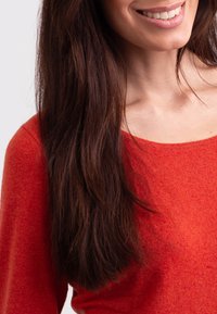 Röd stickad tröja med rund halsringning och långa ärmar. Tyget ser mjukt ut med en texturerad yta, som delvis täcker mörkbrunt hår.