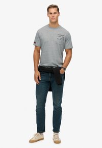 Grå kortærmet t-shirt med en brystlomme og logo, parret med mørkeblue jeans og beige sneakers. Sort striktrøje knyttet om taljen.