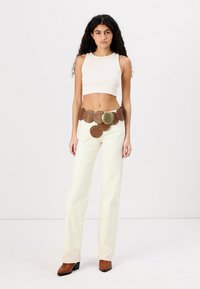 Witte crop top, crème wijde broek en een grote bruine riem met cirkelvormige accenten. Bruine hakken maken de outfit compleet.