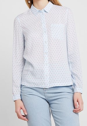 Kvinde iført lyseblå skjorte med lange ærmer, små røde prikker og brystlomme, kombineret med lyseblå jeans.