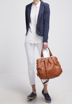 Sac fourre-tout en cuir marron avec design à rabat, quincaillerie argentée et poignées doubles ; associé à un blazer bleu marine et une tenue blanche.