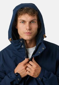 Navyblauwe waterdichte jas met een fleece-voering in de kap, ritssluiting aan de voorkant en drukknoopsluiting. Handen die de jas bij de kraag vasthouden.