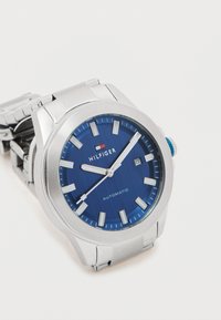Ezüst automata óra kék texturált számlappal, fehér óratartókkal és dátumkijelzővel. Rozsdamentes acél szíjjal és kék korona található.