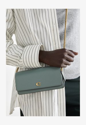 Grønn lær crossover-veske med et gullsymbol, med en strukturert design, kjedestropp og flat rektangulær form.