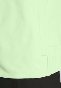 Maillot de sport vert clair avec un motif texturé, un détail de couture latérale et un logo discret à l'ourlet. Le matériau semble lisse et flexible.