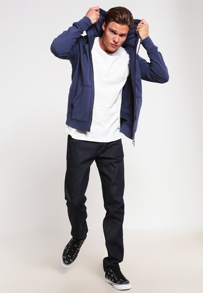 Marineblå hettegenser med glidelås over en hvit t-skjorte, kombinert med mørk denim jeans og svarte joggesko med hvite såler.