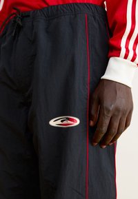 Sorte træningsbukser med røde accenter, elastisk talje og logo på siden. Teksturen ser let og glat ud, velegnet til sportstøj.