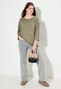 Olivgrünes, langärmliges Oberteil mit einer Tasche, kombiniert mit hellgrauen, weit geschnittenen Jeans und einer kleinen, geflochtenen Handtasche mit schwarzem Boden.