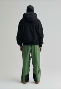 Schwarze Bomberjacke mit Kapuze, kombiniert mit grünen Hosen mit schwarzen Akzenten. Glatte Textur und lockerer Schnitt für Komfort und Bewegungsfreiheit.