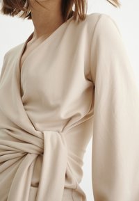 Top enveloppant beige à manches longues, avec un détail noué à la taille et une texture douce et lisse. Design minimaliste.