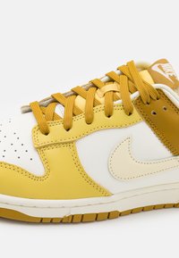 Sneakers Nike z białą skórzaną podstawą i żółtymi akcentami, z teksturowanymi nakładkami, okrągłymi sznurowadłami i brązową metką. Dziurkowany detal w przedniej części.
