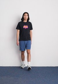 T-shirt noir avec impression graphique multicolore, associé à un short bleu. La personne porte des chaussures de sport blanches. Fond neutre.