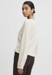 Pull en tricot couleur crème avec un motif en diamant texturé, manches longues et coupe décontractée, associé à un pantalon marron foncé.