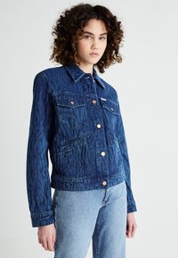 Chaqueta de mezclilla azul oscuro con patrones en relieve, dos bolsillos en el pecho, botones de cobre y un cuello. Combinada con jeans de azul claro.