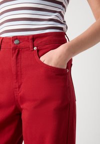 Punased denim püksid sileda tekstuuriga, esi-tasku, nähtav õmblus ja bränditud metallist nööp vööl.
